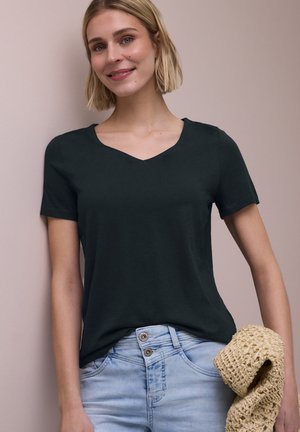 Uśmiechnięta kobieta z krótkimi blond włosami nosi czarny t-shirt z dekoltem w serek oraz jasnoniebieskie, dżinsowe spodnie z wysokim stanem, trzymając plecioną beżową torbę.