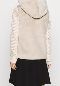 Faux fur-väst i ljus beige med en texturerad stickad ärm, mjuk huva och avslappnad passform, kombinerad med en svart kjol.