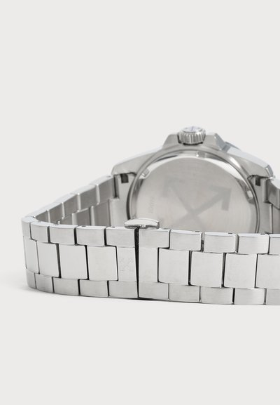 Dos d’une montre-bracelet argentée avec un bracelet métallique à maillons et un fermoir, sur un fond clair.