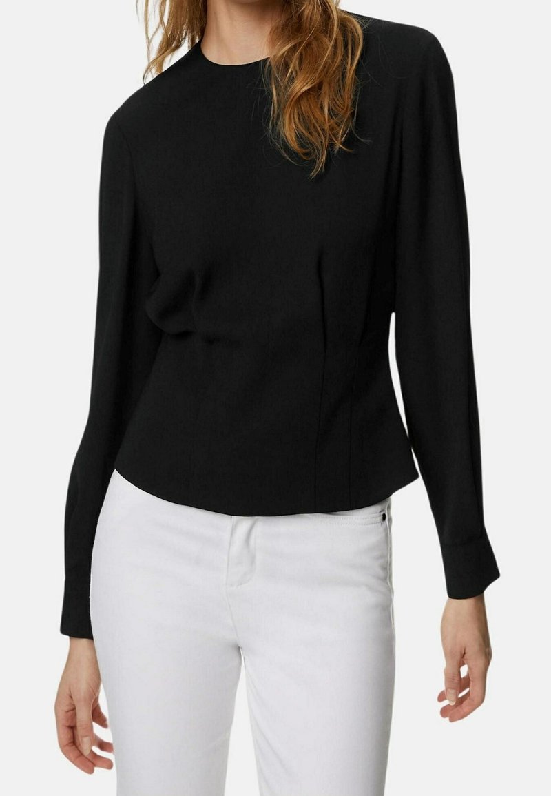 Femme portant un haut noir ajusté à manches longues associé à un pantalon blanc, debout devant un fond uni.