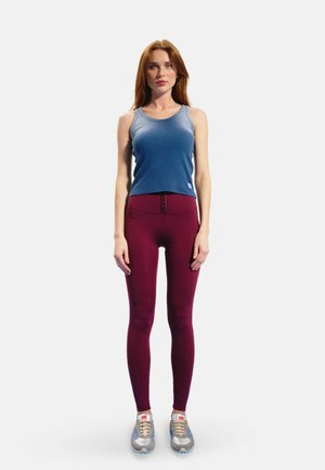 Femme debout face à l’avant portant un haut sans manches bleu, des leggings bordeaux et des baskets grises sur un fond clair uni.