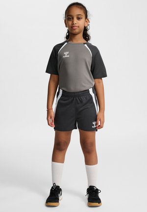 Jong meisje staat met het gezicht naar voren gekeerd, gekleed in een grijs-zwart sportoutfit met witte sokken en zwarte sneakers op een effen achtergrond.