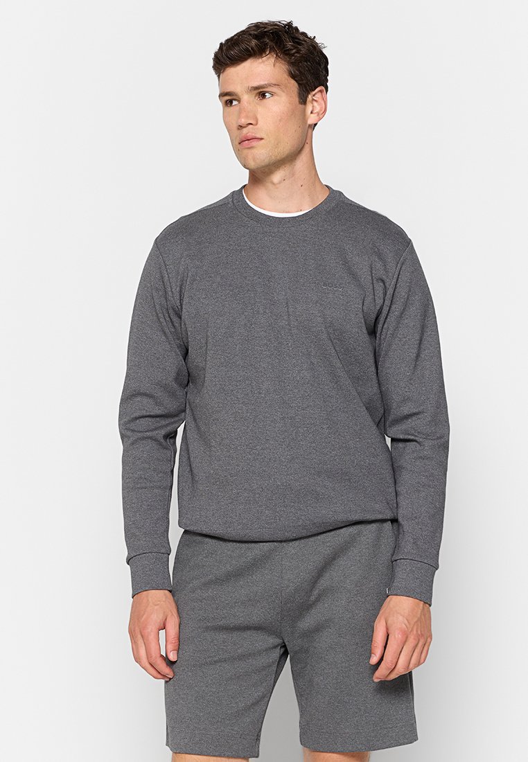 Hombre vestido con una sudadera de manga larga gris oscuro y pantalones cortos a juego, mirando ligeramente hacia la izquierda contra un fondo simple.