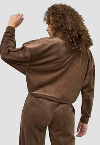 Ruda nubuko medžiagos oversized viršutinė su ilgomis rankovėmis, pasižyminti laisvu siluetu ir matomomis siūlėmis. Modelis vaizduojamas nugarine.