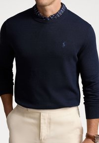 Marinblå stickad tröja med rund hals och långa ärmar, med en subtil logotyp på bröstet; ihop med en prickig skjorta under.