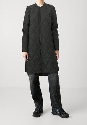 Personne portant un long manteau matelassé noir avec une fermeture éclair à l'avant, un jean sombre délavé et des baskets noires, debout devant un fond clair uni.