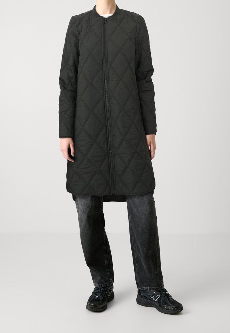 Personne portant un long manteau matelassé noir avec une fermeture éclair à l'avant, un jean sombre délavé et des baskets noires, debout devant un fond clair uni.