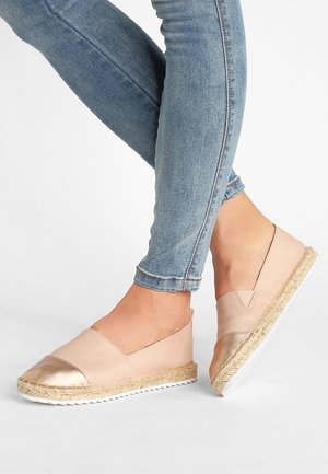 Benen in strakke blauwe jeans die lichtroze instapschoenen dragen met metalen neuzen en geweven zolen op een witte achtergrond.