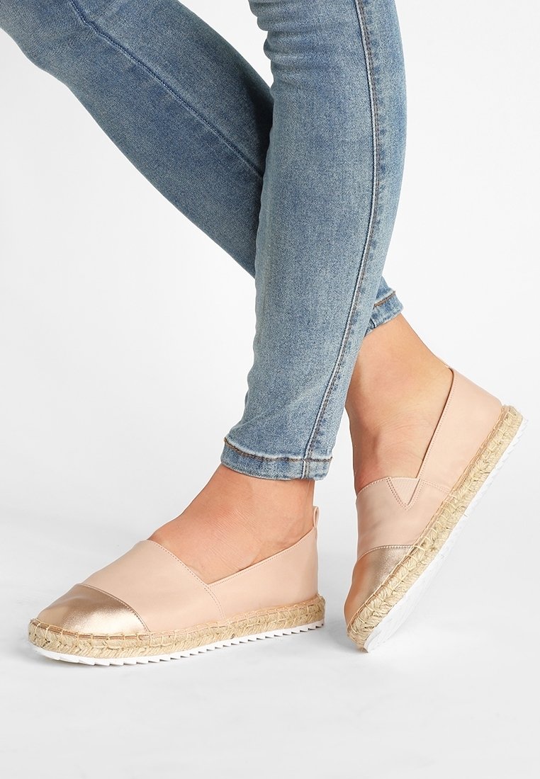 Jambes en jean bleu moulant portant des chaussures slip-on rose clair avec embouts métalliques et semelles tissées sur fond blanc.
