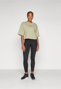 Olivgrünes, kurzes T-Shirt mit kurzen Ärmeln und einem runden Logo, kombiniert mit schwarzen Leggings mit seitlichen Nähten und schwarzen Sportschuhen.