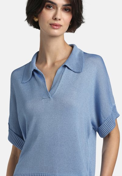 Femeie purtând un tricou polo tricotat cu mâneci scurte de culoare albastru deschis, cu mansete suflecate și cercei aurii, pe un fundal simplu.