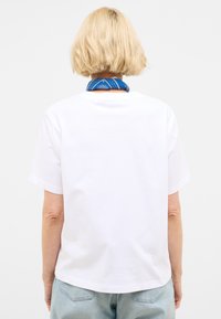 Balta medvilnė t-shirt su apvalia iškirpte ir trumpomis rankovėmis. Galinėje pusėje matyti lygus paviršius be grafinių dizainų, derinama su mėlynu šaliku.