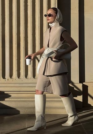 Beige skräddarsydd väst och shortsoutfit, vit bälte och handväska, höga vita stövlar med blixtlåsdetalj, mörka solglasögon och en kaffekopp.