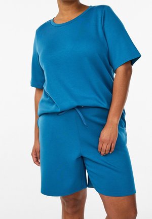 Personne portant un t-shirt bleu à manches courtes et un short bleu assorti avec une taille à cordon, sur un fond neutre.