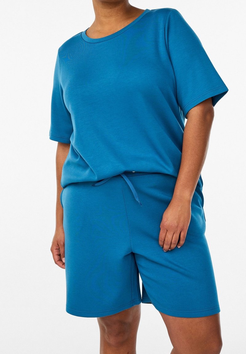 Personne portant un t-shirt bleu à manches courtes et un short bleu assorti avec une taille à cordon, sur un fond neutre.
