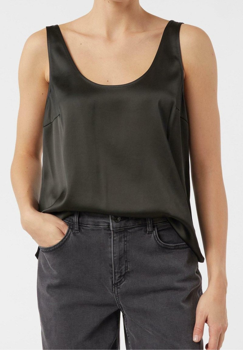Femme portant un top sans manches en satin vert foncé rentré dans un jean taille haute gris foncé, avec une main dans la poche.