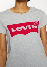 Šedé bavlněné tričko s velkým červeným logem Levi's uprostřed, s klasickým střihem a krátkými rukávy. Měkká textura, bez dalších vzorů.
