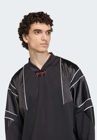 Crni dres od mrežastog materijala, s V-izrezom, bijelim prugama na rukavima i crvenim adidas logotipom na ovratniku. Vidljiv je detalj teksturirane površine.