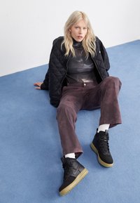 Blond kvinna som sitter på blå matta, iklädd svart vadderad jacka, batikmönstrad polotröja, bruna byxor, vita strumpor och svarta hög-top sneakers.