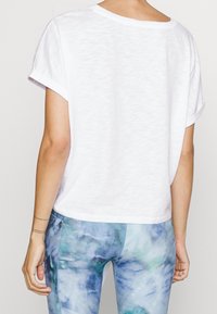 Camiseta blanca de manga corta con un corte holgado y puños doblados, combinada con leggings azules y verdes con un patrón que muestra un efecto teñido anudado.