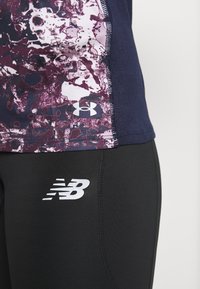 Dopasowany sportowy top z fioletowym wzorem rozbryzgów i granatowymi bokami, zestawiony z czarnymi legginsami z białym logo New Balance.