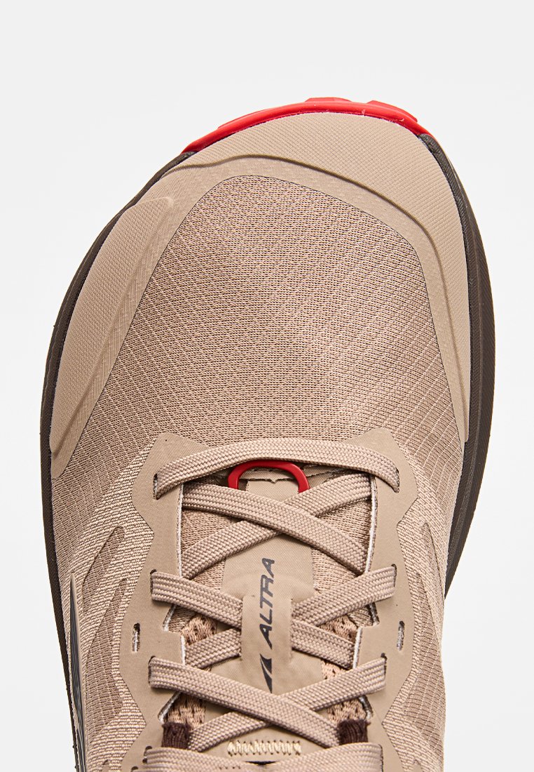 Zapatilla deportiva beige con parte superior de malla, detalles en rojo y suela negra. Presenta una puntera reforzada y una superficie texturizada. Cierre con cordones.