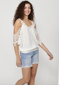 Top bianco a spalle scoperte con dettagli in pizzo e una consistenza morbida, abbinato a pantaloncini di jeans blu chiaro con ricami floreali e orli a scallop.