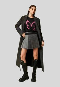 Cappotto lungo a quadri grigi, t-shirt nera con grafica di una farfalla rosa, gonna grigia a scacchi con zip laterale e stivalli neri con lacci.