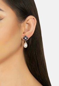 Gold earrings featuring a blue gemstone floral design with a pearl drop. The earrings are worn on an ear with straight, dark hair.