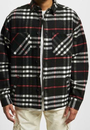 Mężczyzna ubrany w czarno-biało-czerwoną koszulę w kratę zapinaną na guziki, na białej koszulce, do tego jasne spodnie, z rękami swobodnie opuszczonymi wzdłuż ciała.