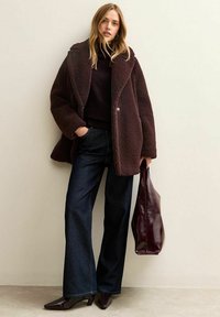 Donna che indossa un cappotto teddy marrone scuro, un dolcevita nero, jeans a gamba larga e stivaletti con punta, mentre tiene una grande borsa tote bordeaux.