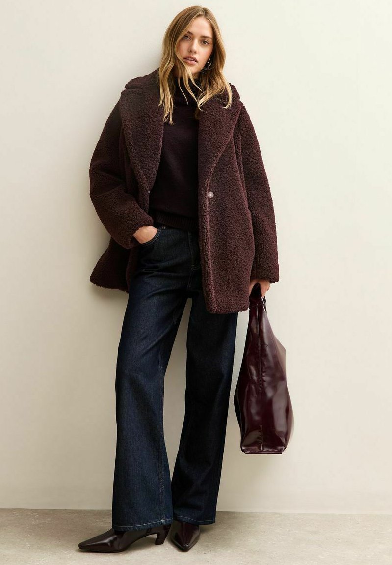 Donna che indossa un cappotto teddy marrone scuro, un dolcevita nero, jeans a gamba larga e stivaletti con punta, mentre tiene una grande borsa tote bordeaux.