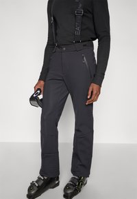 Μαύροι παντελόνες για σκι με ρυθμιζόμενες τιράντες, τσέπες με φερμουάρ, ριμπ κοψίματα στα πόδια και εφαρμοστή γραμμή. Συνδυάζονται με μαύρα παπούτσια για σκι.