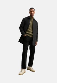 Zwarte jas over een olijfgroene gebreide trui, zwarte broek en beige sneakers. De jas heeft knopen en een recht gesneden ontwerp.