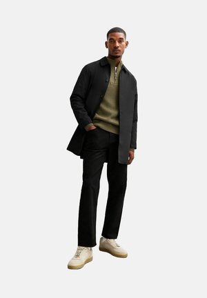 Abrigo negro sobre un suéter de punto verde oliva, pantalones negros y zapatillas beige. El abrigo tiene botones y un diseño de corte recto.