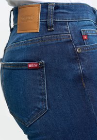 Prim-plan al buzunarului din spate al blugilor denim albastri, cu etichetă maro din piele Big Star și etichete de țesătură roșie pe buzunare.