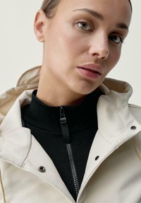 Béžová mikina s kapucňou cez čierny top s vysokým golierom, ktorý má zips. Hladká textúra s viditeľným prešívaním a detailmi na cvočky.