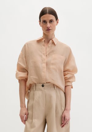 Frau mit zurückgekämmtem Haar, die ein lockeres, pfirsichfarbenes Hemd mit Knöpfen und hochtaillierte beige Hose trägt, vor einem schlichten weißen Hintergrund stehend.