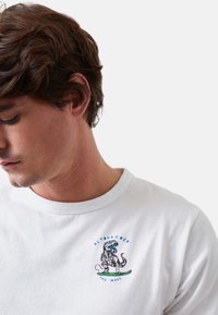 Camiseta de algodón blanca con un gráfico colorido de dinosaurio y el texto "ALTONADOCK THE WAVE", cuello redondo y mangas cortas.