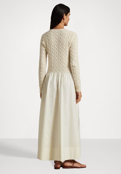 Mulher vestindo um suéter de malha tipo cable em creme, combinado com uma saia longa e fluida em off-white e sandálias rasas marrons, de pé, de costas.