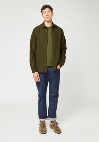 Olijfgroen knoopshirt over een donkergroene T-shirt, gecombineerd met een blauwe spijkerbroek en bruine veterschoenen. Het shirt heeft een subtiele logo.