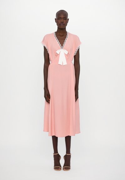 Abito rosa con scollo a V e maniche corte, caratterizzato da un bordo in pizzo bianco. Include un dettaglio con fiocco legato nella parte anteriore e una gonna lunga fino a metà polpaccio, fluida.