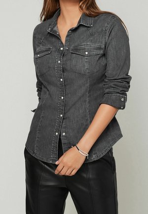 Chemise en denim gris foncé à manches longues, boutons sur le devant, poches poitrine et poignets retroussés. Texture lisse avec des accents de boutons en métal.