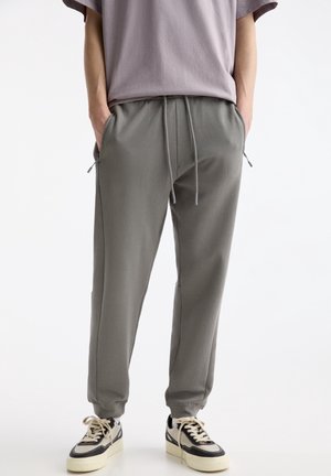 Person trägt graue Jogginghose mit Kordelzug und Reißverschlusstaschen sowie weiße Sneakers mit schwarzen Akzenten, steht mit den Händen in den Taschen.