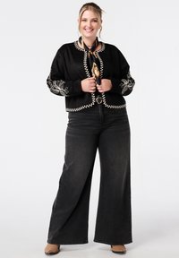 Glunderende vrouw in zwart geborduurd jasje, met een gemusterd sjaal, wijde zwarte spijkerbroek en bruine schoenen, staand voor een witte achtergrond.