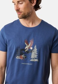 Blå kortärmad t-shirt i bomull med ett motiv av en örn, träd och vatten. Texten "SWEDEMOUNT" tryckt under designen.