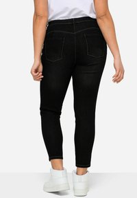 Vaqueros negros de talle alto con una forma ajustada y dos bolsillos traseros. La tela tiene una textura suave y un brillo sutil. Se llevan con zapatillas blancas.