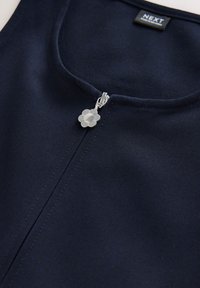 Tumesinist riietustoonikat ümmarguse kaelusega ja lukuga, millel on hõbedast lillekujulise tõmblukukäepide. Kangas on tekstuuriga ja tugev.