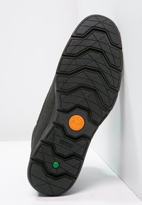 Semelle de chaussure noire avec des rainures profondes, logo Timberland orange et petit détail hexagonal vert sur fond blanc.