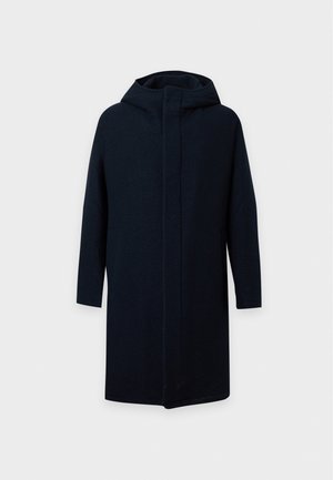Un homme pose en portant un long manteau bleu marine, un col roulé côtelé couleur rouille, un pantalon foncé et des mocassins noirs, sur un fond blanc uni.